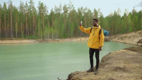 Un hombre negro con una mochila usa un teléfono móvil mientras camina por el bosque en las montañas al otro lado del lago. Tomar una foto y blog - Metraje, vídeo
