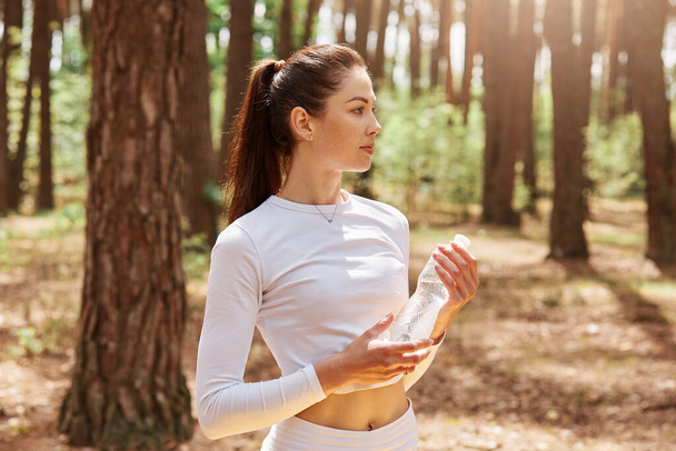 Fitness beautiful woman with dark hair and ponytail holding bottle of water and looking away, posing after exercising in forest. Female athlete in white top work out outdoor. - Photo, image