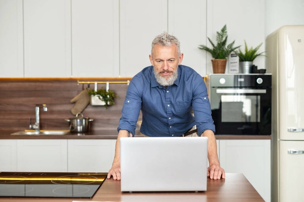 Senior man freelancer stands near countertop in the kitchen and staring at the laptop - Foto, imagen