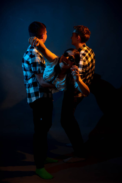 Two guys in a plaid shirt are holding a girl on a dark background in a studio with multicolored light - Foto, Imagen