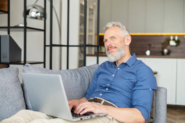 Senior male freelancer lying down on the sofa and using laptop - Фото, изображение