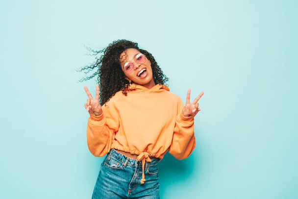 Beautiful black woman with afro curls hairstyle.Smiling model in orange hoodie and trendy jeans clothes. Sexy carefree female posing near blue wall in studio in sunglasses.Shows peace sign - Фото, зображення