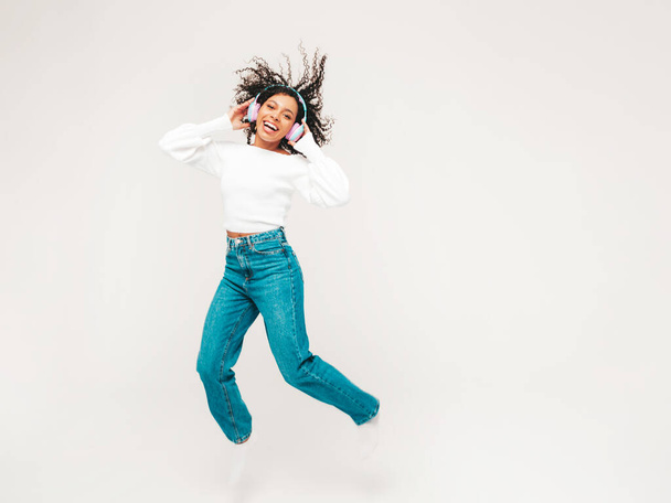 Beautiful black woman with afro curls hairstyle.Smiling model in sweater and jeans.Sexy carefree female listening music in wireless headphones. Jumping in studio on white background.Dynamic movement  - Foto, Bild