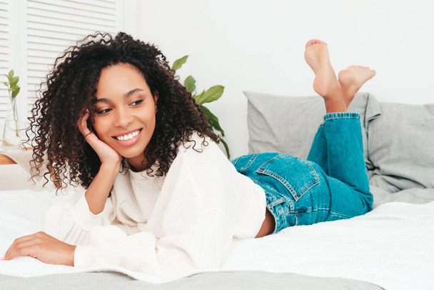 Young beautiful black female with afro curls hairstyle. Sexy smiling model in sweater and trendy jeans clothes lies on bed in posh apartment or hotel room in white interior in the morning - 写真・画像