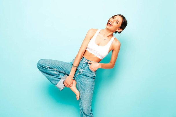 Beautiful smiling woman dressed in white jersey top shirt and jeans. Sexy carefree cheerful model enjoying her morning. Adorable and positive female posing near blue wall in studio - Φωτογραφία, εικόνα