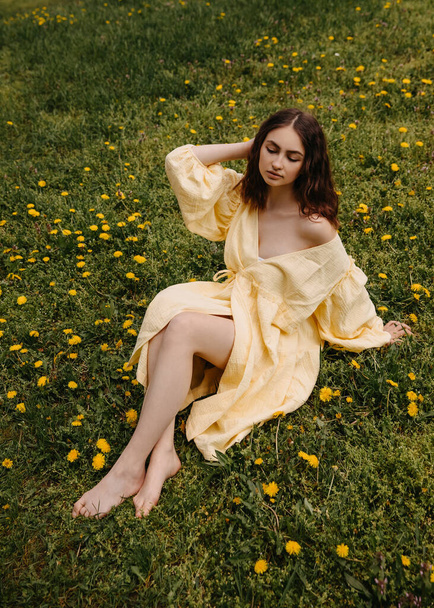 Young barefoot woman in a yellow muslin dress, sitting in a field with green grass and dandelions. - Photo, Image