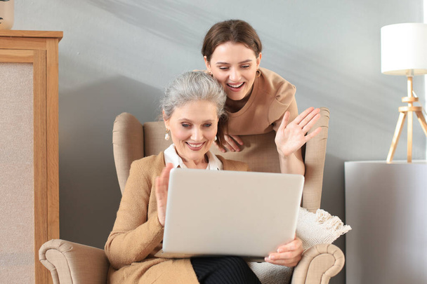 Happy elderly middle mother sitting on chair with her daughter, looking at laptop. Young woman showing video, photos to mommy, trusted relations. Family concept - Fotoğraf, Görsel