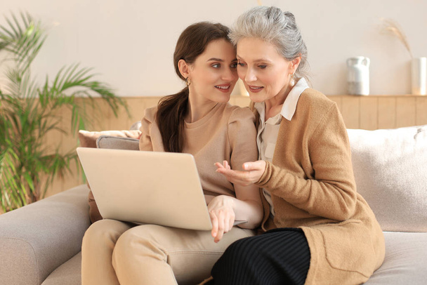 Happy elderly middle mother sitting on couch with her daughter, looking at laptop. Young woman showing video, photos to mommy, trusted relations. Family concept - Fotografie, Obrázek