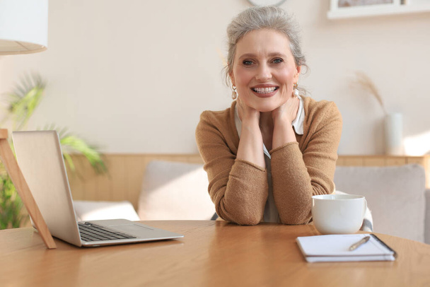 Middle aged woman sitting at a table with a laptop and looking at the camera smiling - Foto, Imagem