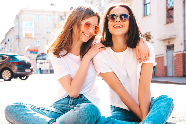 Two young beautiful smiling hipster female in trendy summer white t-shirt clothes and jeans.Sexy carefree women posing on the street background. Positive models having fun, hugging and going crazy - Foto, Bild