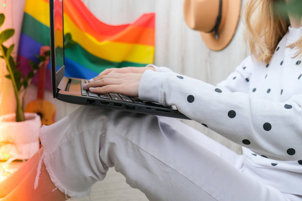 Jonge millennial hippie vrouw zit op het balkon met behulp van laptop. LGBTQ regenboogvlag op achtergrond. Online onderwijs onderwijs. Werk vanuit huis. Modern kantoor. - Foto, afbeelding