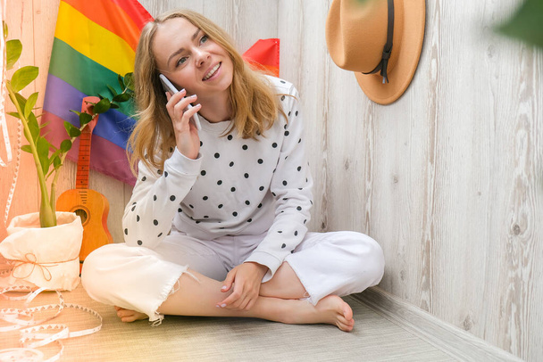 Young millennial hippie woman sitting on balcony using mobile phone. LGBTQ rainbow flag on background. Phone texting, video call, taking selfie. Blogger. Work from home. - Foto, Bild