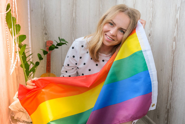 Joven mujer hippie rubia milenaria con bandera LGBTQ arco iris en el balcón de casa. Paz y libertad el símbolo de la comunidad LGBT, pasar y compartir el tiempo amoroso. Apoyo a la comunidad LGBTQ. - Foto, imagen
