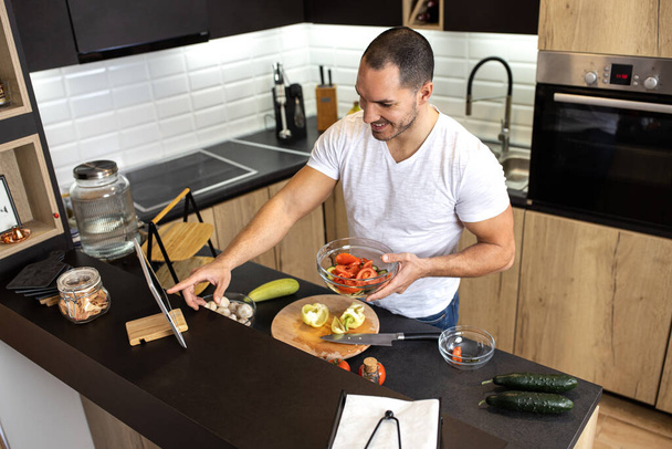 Young adult man holding a bowl of fresh vegetables in the kitchen of his apartment as he prepares a dish - Foto, immagini