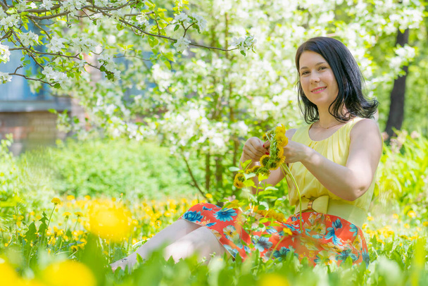 明るい晴れた日。明るい夏のドレスを着た若い女性が緑の芝生の上に座って、黄色のタンポポの花の花輪を織ります。屋外レクリエーションと素晴らしい気分. - 写真・画像