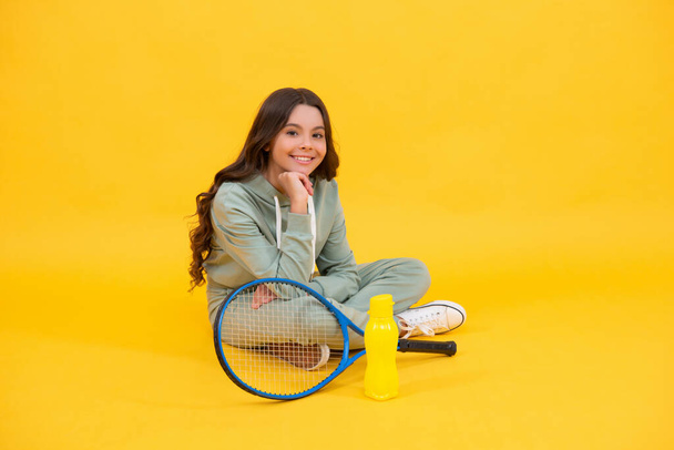 vrolijk kind zitten in sportkleding met badminton racket en water fles op gele achtergrond, ontspannen - Foto, afbeelding
