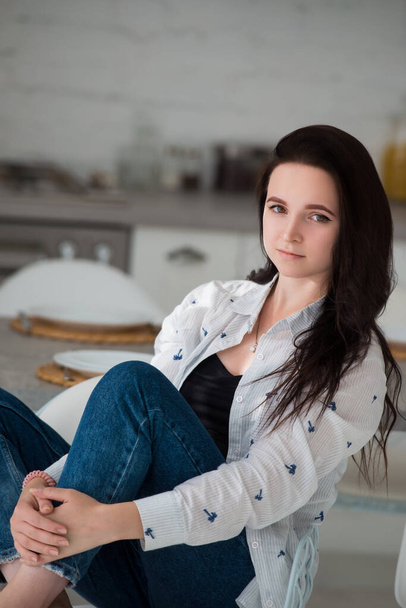 Young brunette girl student sits on chair in the kitchen, hugging her knees with her hands. Woman dressed in casual clothes jeans and shirt - Foto, Imagen