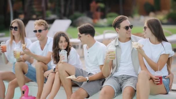 Cute caucasian woman feeding yummy pizza to her boyfriend on the background of their friends near poolside. Young people happily spending quality time together while eating cheese pizza on party - Footage, Video