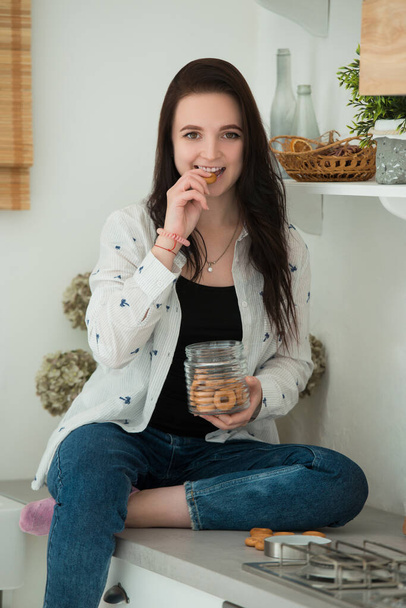 Smiling young brunette woman is sitting at the table in the kitchen. The girl eats bagels and cookies. A dry snack is not a great weight loss idea. - Foto, afbeelding