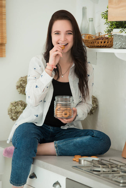 Smiling young brunette woman is sitting at the table in the kitchen. The girl eats bagels and cookies. A dry snack is not a great weight loss idea. - Foto, Imagem
