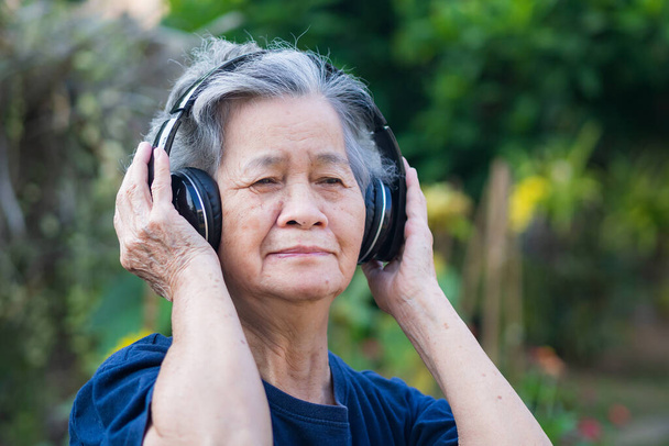 Seniorin mit drahtlosem Kopfhörer hört im Garten ein Lieblingslied. Asiatische alte Frau, kurze weiße Haare genießen die Musik. Konzept der Alten- und Krankenpflege - Foto, Bild