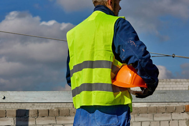 Professionele bouwer in werkkleding houdt een oranje harde hoed vast terwijl hij op het dak staat van een gebouw in aanbouw en wegkijkt. Blauwe lucht - Foto, afbeelding