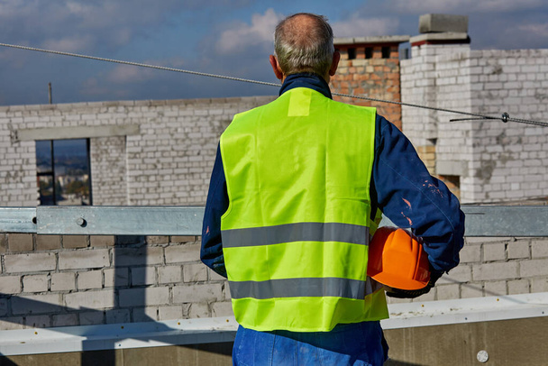 Az érett munkás védőruhában egy narancssárga biztonsági sisakot tart, miközben az építés alatt álló épület tetején áll. Kék ég. - Fotó, kép