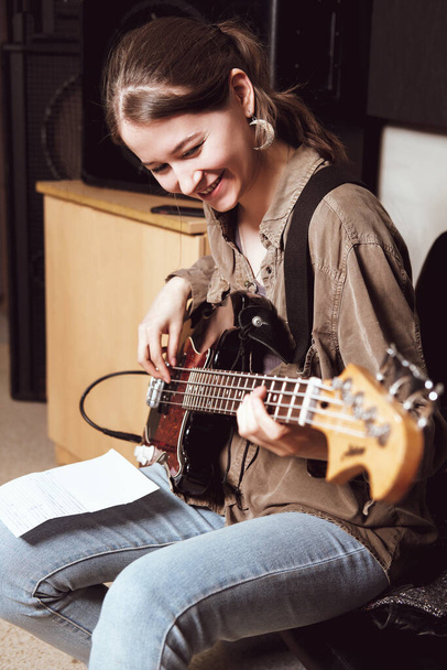 mladá usměvavá žena hrající na baskytaru a čtoucí akordy na zkoušce kapely. svislá velikost - Fotografie, Obrázek
