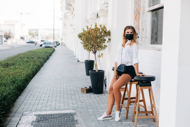 молода жінка в захисній масці чекає на своє замовлення, сидячи на стільці у відкритому кафе під час пандемії. Соціальна відстань, захист
 - Фото, зображення