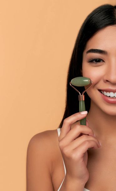 A young Asian woman with clean radiant skin makes a facial massage with a quartz roller on a beige background. Spa care, facial skin care, beauty cosmetology. - Foto, immagini