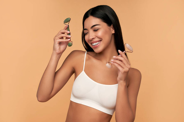 Una joven asiática con la piel radiante limpia hace un masaje facial con un rodillo de cuarzo sobre un fondo beige. Cuidado del spa, cuidado de la piel facial, cosmetología de belleza. - Foto, Imagen