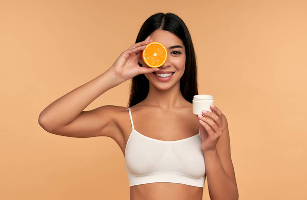 Retrato de niña asiática sosteniendo crema facial anaranjada e hidratante aislada sobre fondo beige. Concepto de cuidado facial de la piel, cosmética natural. - Foto, imagen