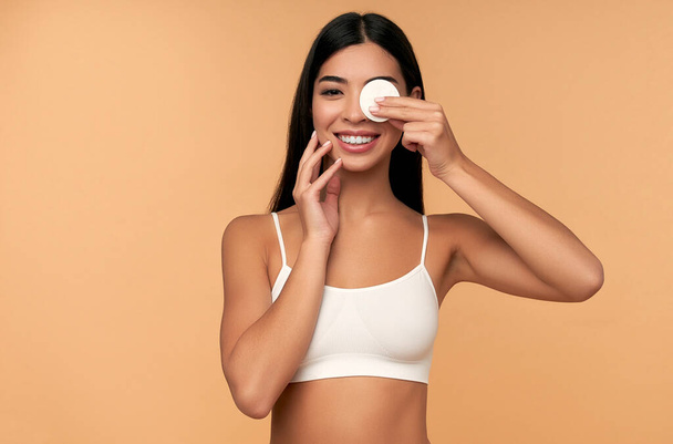 Young woman cleans beauty face using cotton pads, cleansing lotion and face toner to remove makeup. Nice girl with beautiful skin. Skin care concept. Beige background, copy space - Photo, Image