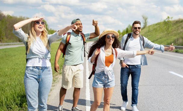 Happy multiethnic friends trying to get free ride, hitchhiking on highway, showing thumb up gestures outdoors, panorama - Фото, изображение