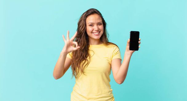 young pretty woman feeling happy, showing approval with okay gesture and holding a smart phone - Foto, afbeelding