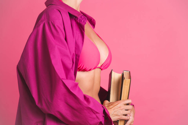 Beautiful fit tanned sporty woman in bikini and shirt holding book go to beach to read on pink background - Photo, Image