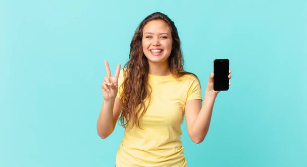 young pretty woman smiling and looking friendly, showing number two and holding a smart phone - Foto, afbeelding