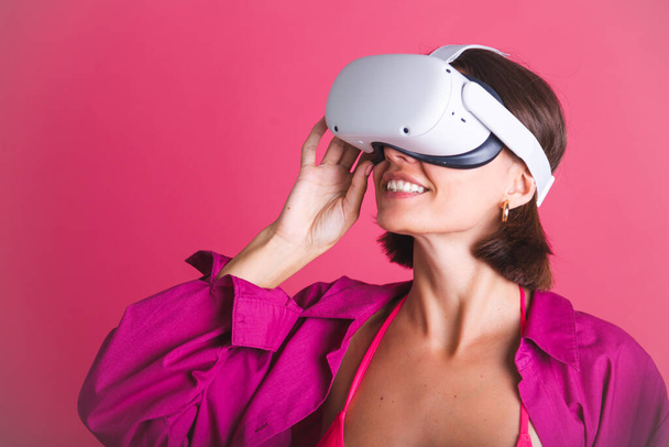 Beautiful fit tanned sporty woman in bikini and shirt in virtual reality glasses on pink background - Fotoğraf, Görsel