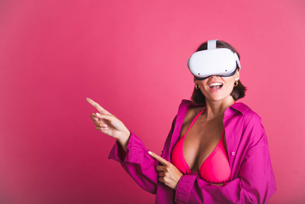 Beautiful fit tanned sporty woman in bikini and shirt in virtual reality glasses on pink background point fingers left - Valokuva, kuva