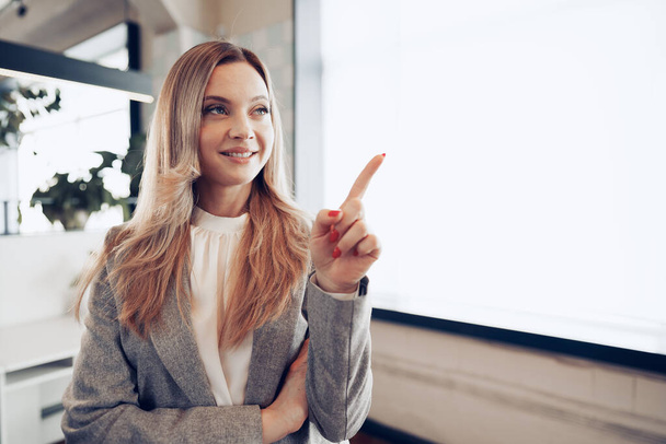 Young beautiful woman standing near window in office and pointing finger to copy space - Foto, imagen