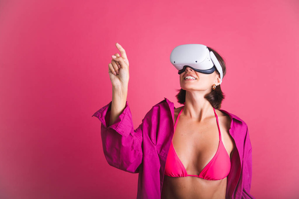 Beautiful fit tanned sporty woman in bikini and shirt in virtual reality glasses on pink background click finger left - Foto, Imagem