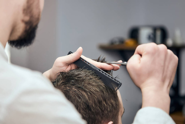 Профессиональный парикмахер делает стрижку молодому человеку, используя ножницы и расческу в парикмахерской. Крупный план, избирательный фокус - Фото, изображение