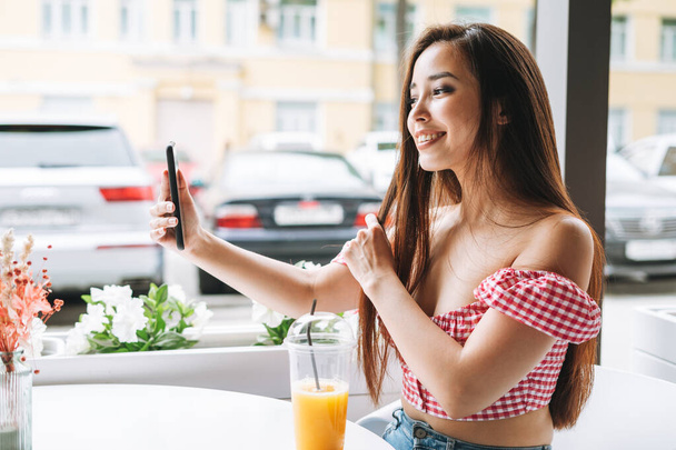Uśmiechnięta piękna azjatycka młoda kobieta z długimi włosami grabiąca selfie na telefonie komórkowym na tarasie letniej kawiarni. Dziewczyna w czerwonych ubraniach na wakacjach w kurorcie - Zdjęcie, obraz