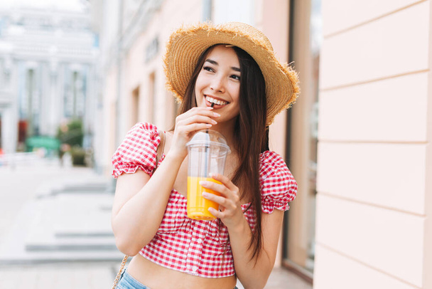 Uzun saçlı, saman şapkalı, gülümseyen Asyalı genç bir kadın. Şehir caddesinde portakal suyu içiyor. - Fotoğraf, Görsel