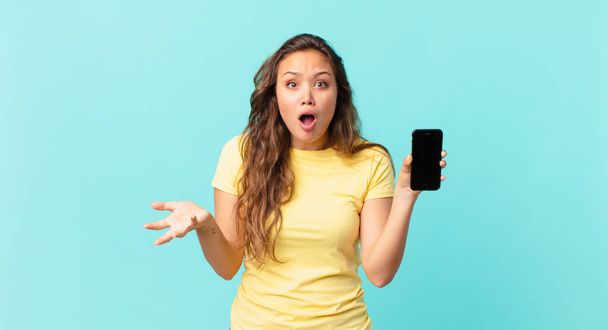 young pretty woman feeling extremely shocked and surprised and holding a smart phone - Foto, afbeelding