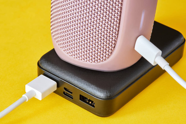 Power bank charges portable audio speaker using a USB cable on a yellow background. Close-up, selective focus - Photo, Image