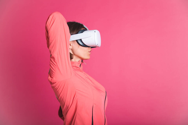 Young fit woman in sport wear jacket and virtual reality glasses stretching herself on pink background - Photo, Image
