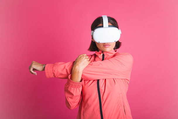 Young fit woman in sport wear jacket and virtual reality glasses stretching herself on pink background - Fotó, kép
