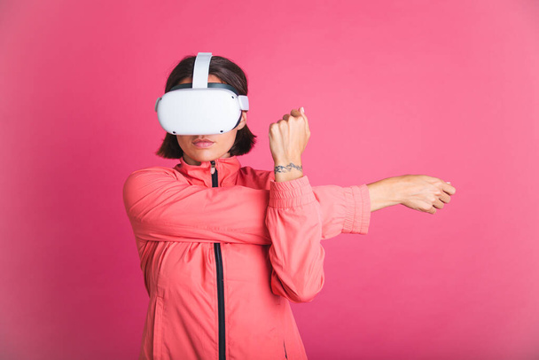 Young fit woman in sport wear jacket and virtual reality glasses stretching herself on pink background - Фото, изображение