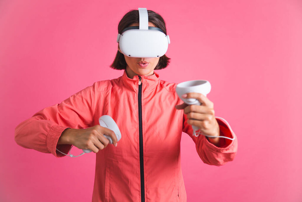Young fit woman in sport wear jacket and virtual reality glasses playing boxing fight games on pink background - Photo, Image